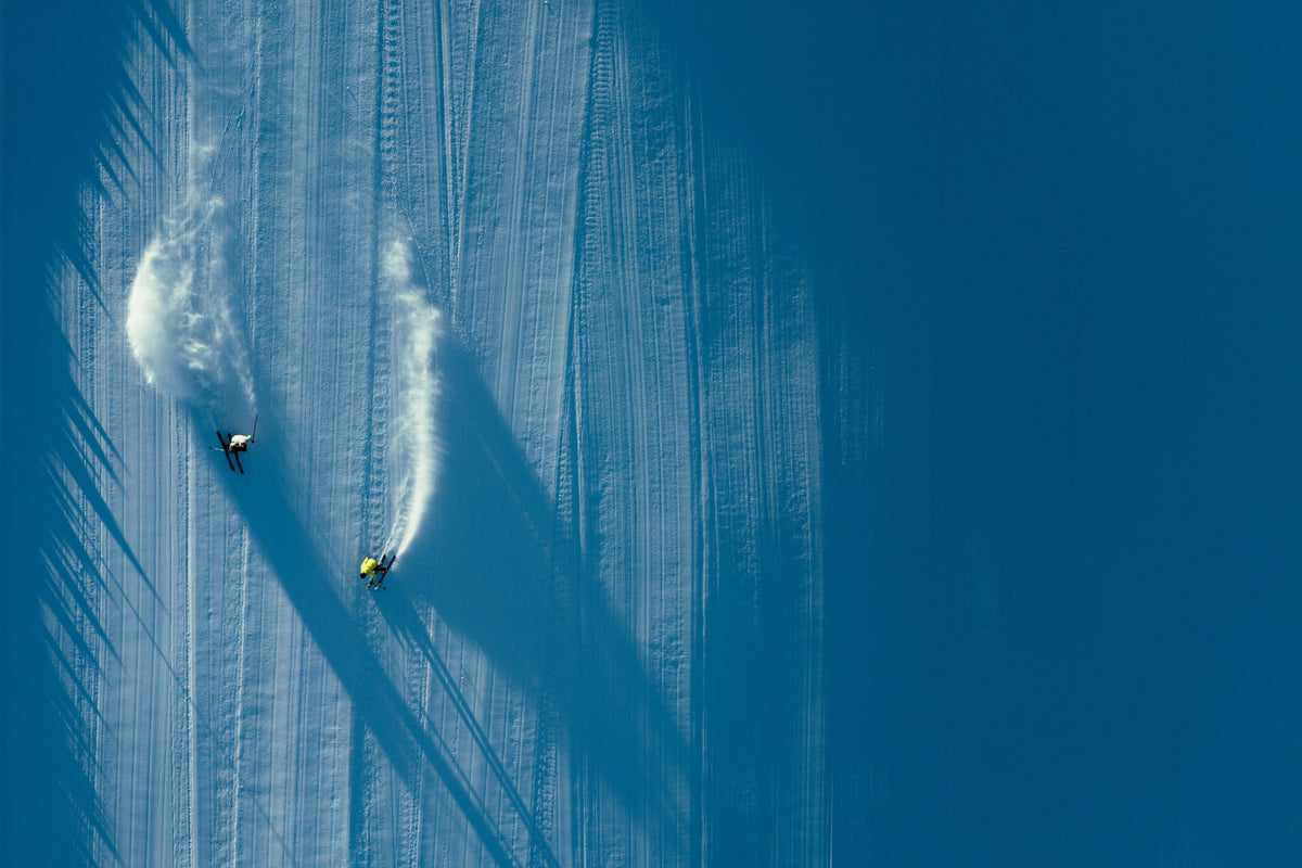 Two skiers carving turns down a groomed slope, seen from above